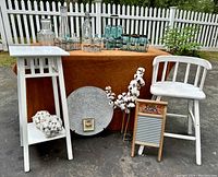 Wide view of lot showing white spindle-backed high stool, mission style white corner table, various glass jars and bottles on table covered with brown cloth, cotton stems, decorative plate and washboard on ground in front.