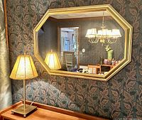 Photo of the octagonal gold framed wall mirror hung on a dark floral wallpapered wall above a wooden surface with a lamp beside it.
