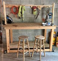 Wide front view of the pine workbench showing two wooden stools, overhead dowel bar, hanging ropes, chains, extension cord, hand saw, bar clamp, Ryobi miter saw on the right side of the bench.