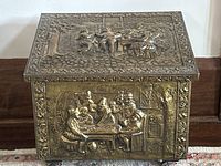 Front view of the antique brass fireplace box showing embossed social scene on the lid and side.