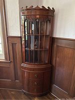 Full frontal view of antique mahogany corner china cabinet showing curved glass door with wood mullions, carved upper trim, and base drawers with brass pulls.