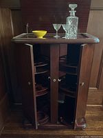 Front view of antique mahogany bar cabinet with doors open showing circular rotating shelves inside for storing bottles and glasses, glass top has a crystal decanter and glasses on it.