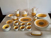 Full set of vintage mid-century dishware with sunflower motif arranged on table showing plates, bowls, cups, saucers, pitchers, coffee pot, and butter dish.
