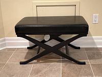 Front angle view of black leather bench with wooden X-frame base showing padded seat and legs on tile floor