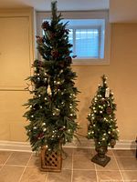 Pair of pre-lit artificial Christmas trees shown side by side against a wall with a small window. Larger tree is in a wooden crate base, decorated with pinecones and red berries, illuminated with warm white lights. Smaller tree has similar decorations in an urn-style base.