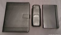 Set of three black leather items: large business portfolio with magnetic clasp on left, 3-count cigar holder in middle, and telephone/address book on right with elastic band closure.