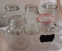 Front view of 5 glass canisters showing clear glass bodies and silver metal clasp lids, one with black chalkboard label and one with red silicone seal.