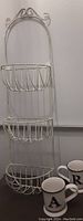 Full view of white iron three-basket wall shelf with decorative curled top showing all three baskets and partially visible mugs below.
