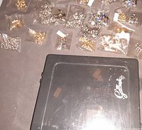 Photo showing multiple small plastic bags of rhinestones in various colors and sizes spread over a surface with a clear plastic box lid labeled 'Ge Magic'.