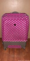 Front view of pink polka dot large hard shelled suitcase with visible front zippered pocket and carry handle on top.