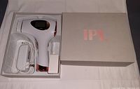 White handheld IPL hair removal device with rose gold accents inside original gray box with 'IPL' lettering. Includes power adapter and replacement flash window.