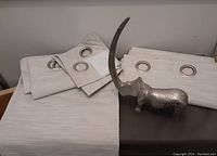 Set of folded light beige/gray blackout curtains with visible metal grommets beside metallic rhino sculpture with long protruding tusk laid on surface.