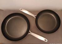 Top view of two frying pans, one larger (31 cm) and one smaller (27 cm), with black non-stick interiors and metal handles.
