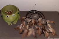 Green ceramic frog planter next to large dream catcher with three rings, feathers, beads, and metal wind chimes.