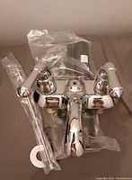 Photo showing the chrome vanity faucet laid flat with attached handles and mounting hardware still in protective plastic wrapping.