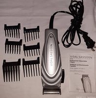 Vidal Sassoon electric hair clipper with 7 black plastic guide combs and original manual showing all included accessories laid out on a fabric background.