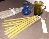 Blue and green crackle glass miniature pitchers/vases, hand dipped yellow taper candles, and beeswax candles bundled in white ribbons on a cloth surface.