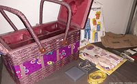 Purple rectangular sewing basket with butterfly decorations and handles, open showing storage compartments, placed on table with some accessories and patterns nearby.