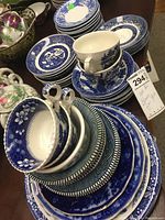 Stack of blue and white patterned plates, teacups and saucers with floral and scenic designs. Various patterns stacked on a table.
