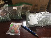 Zoomed out image showing several plastic bags filled with marbles on a table, one smaller bag contains colorful marbles. A ballpoint pen is placed to show scale.