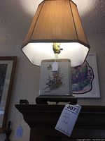 Full view of beige ceramic lamp with floral motif, beige fabric lampshade, sitting on wooden surface with framed art in background.