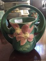 Front view showing the rounded two-handled vase with raised Clematis flower design, green matte glaze with pink and yellow flowers.