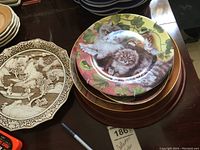 Collector plates with a colorful kitten design and sepia scenic design stacked on a table, with a pen and lot tag visible.