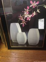 Framed floral art poster with pink orchids and ribbed white vases against a black background.