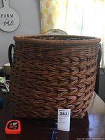 Large brown woven wicker basket with visible handles and thick reinforced rim, placed on a table for scale. A tape measure and MaxSold tag are included for size reference.