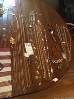 Photo showing 11 assorted long chain necklaces arranged on wooden table, featuring various pendant styles including crosses, sun shapes, disks, and leaf shapes in metal tones.