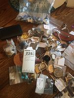 Overview photo showing multiple small bags with assorted costume jewelry and accessories spread on a wooden surface, including buttons, cuff links, tie tacs, belt buckles, and key rings.