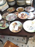 Photo showing 11 decorative plates featuring painted puppies in natural settings, laid out on table
