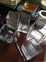 Six square vases of varying materials and designs displayed on a dark wooden surface, including clear glass, clear acrylic, and metal decorative vase.