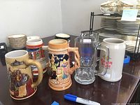 Five large beer steins including German ceramic, glass, and branded examples on table with dishware in background