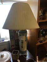 Front view of tall white and blue ceramic lamp with white pleated fabric shade, showing decorative floral pattern and lamp size in room context.