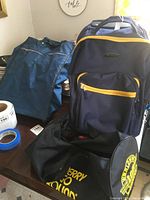 Photo showing three luggage cases on a table: a Rockland branded blue wheeled case with yellow zipper, a plain blue foldable case, and a cylindrical black athletic case with yellow print.