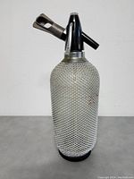 Front view of classic glass soda siphon bottle with white wire mesh covering and black dispensing top against plain background.