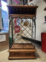 Front view of the wine rack showing metal diamond grid body, wooden top shelf with glass slots, and wooden bottom drawer with handle.