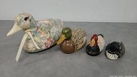 Four decorative bird figurines arranged in a row including largest floral ceramic duck, two smaller ducks, and one small loon.