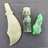 Three jade items displayed on a dark background: a curved jade sword blade, cylindrical jade seal, and a carved bird figure.