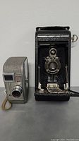 Two vintage cameras placed side by side on a flat surface - Kodak No. 1A Autographic Jr. folding camera with bellows on right and compact silver Keystone Capri 8mm film camera on left.