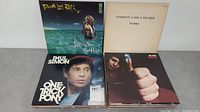 Front view of four LP vinyl albums featuring David Lee Roth, Emerson Lake & Palmer, Paul Simon, and an unknown album with a hand holding a pin.