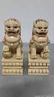 Pair of beige resin Chinese Foo Dog figurines on rectangular brick-like bases, one with paw on a ball and the other with raised paw
