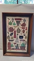 Full view of vintage linen artwork framed in a dark wooden frame showing the colorful images of kitchen utensils and vessels on linen fabric.