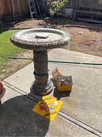 Concrete bird bath standing on concrete patio with two birdhouses and yellow 'SONGBIRD CROSSING' sign on ground around it.