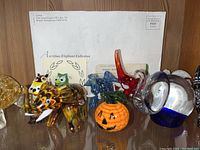 Photo of amber and blue elephants, multicolored owl pair, multicolored bird, and pumpkin figurines arranged on a glass shelf with certificates in background