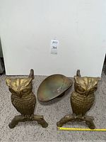 Front view of two brass owl andirons and oval brass hearth scale plate on floor with measuring tape showing approximately 13.5 inches tall for owls.