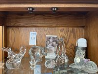 Photo showing multiple clear glass animal sculptures including swan, bear, standing cat, and faceted crystal egg with a small angel figurine.