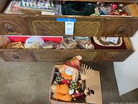 Cardboard box containing a fabric scarecrow figure wearing a green plaid shirt, straw hat with pumpkin applique, and three small orange fabric pumpkin decorations alongside a small hand rake.