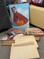 Photo showing framed rocket launch print with a red cape-like shape, newspapers with visible headline 'THEY'RE OFF TO MOON!', and envelopes with handwritten notes about music festivals and historical dates.